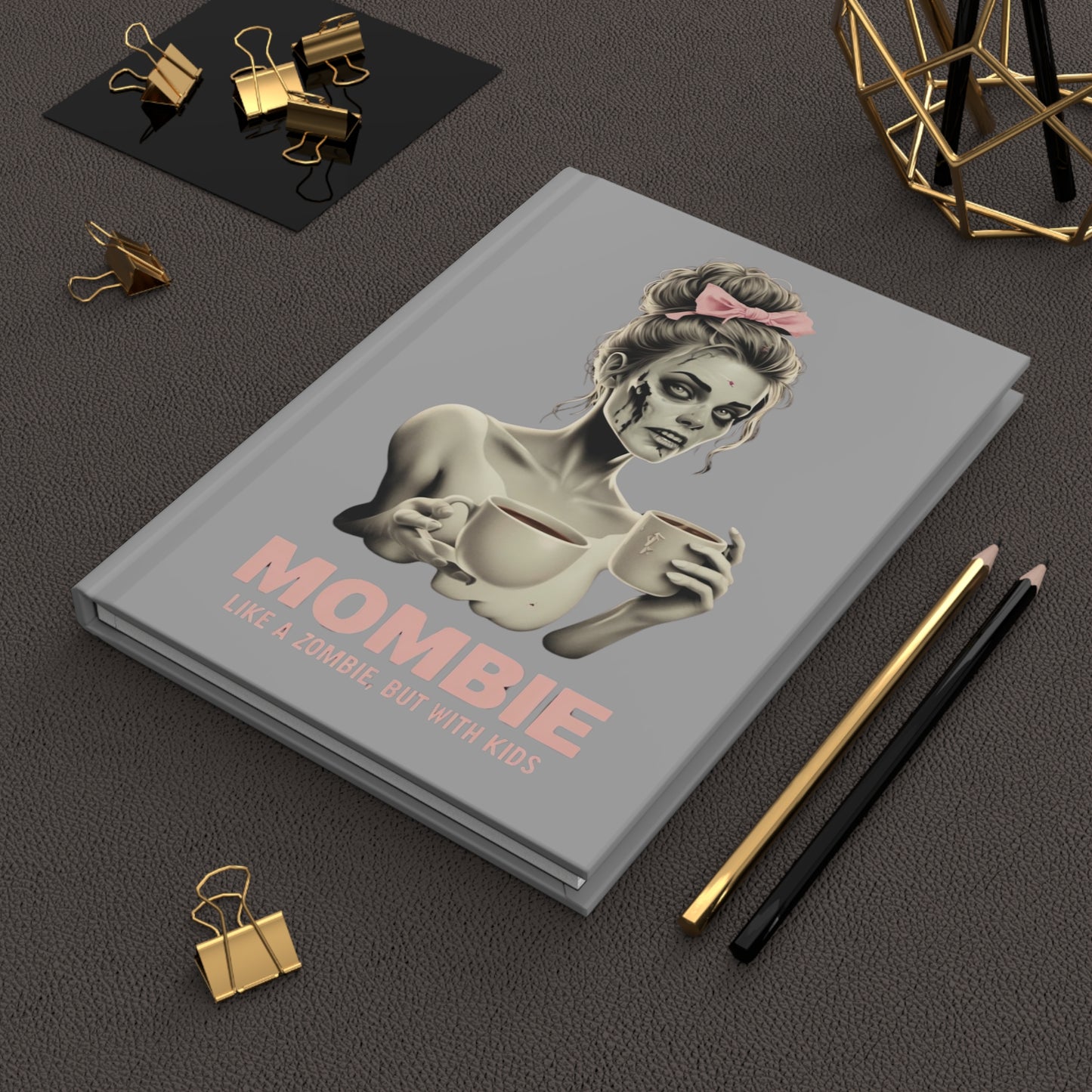 Hardcover journal with a grey cover featuring a cartoon-style zombie mom holding two coffee mugs, wearing a pink bow, with the words "Mombie – Like a Zombie, But With Kids" printed in bold pink font.
