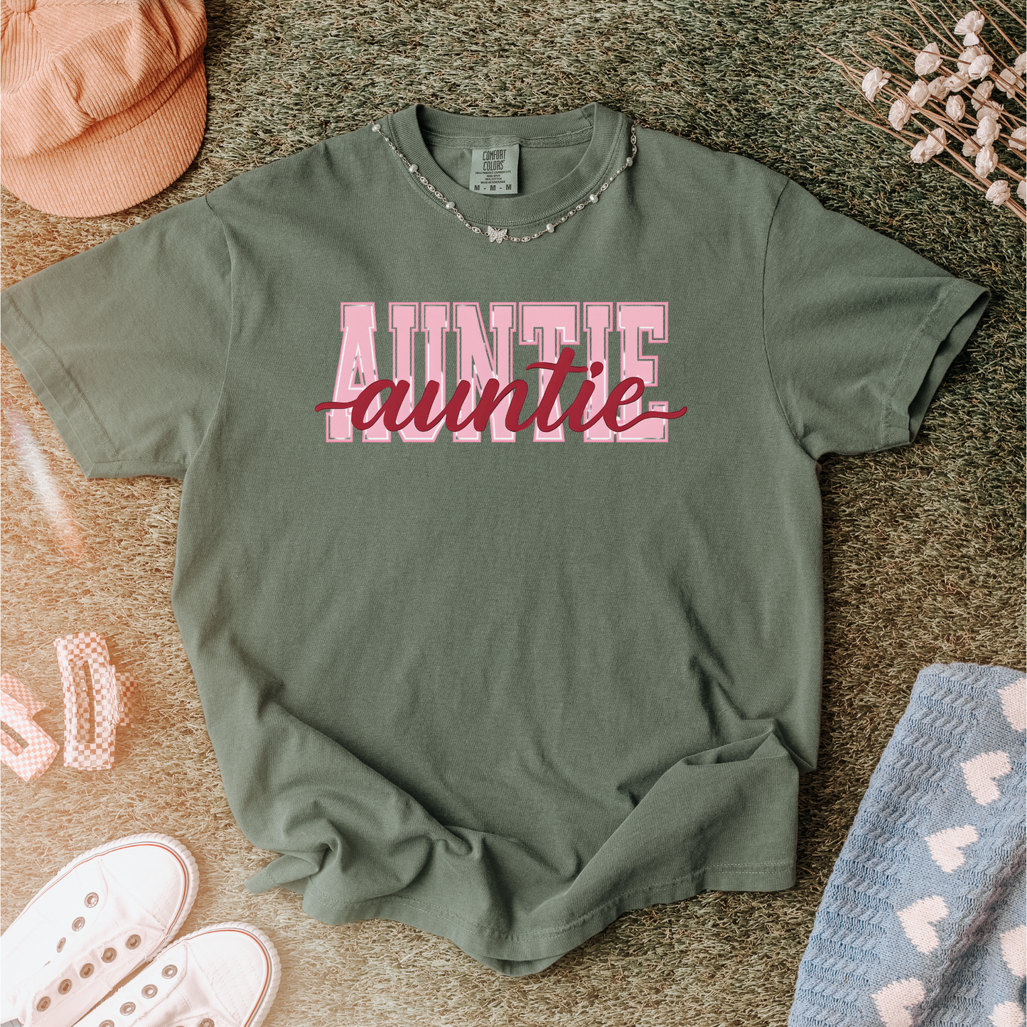 Women wearing graphic t-shirts with the word “AUNTIE” in bold pink varsity letters and a red cursive overlay, available in black, blue, green, and ivory, styled in lifestyle and casual settings