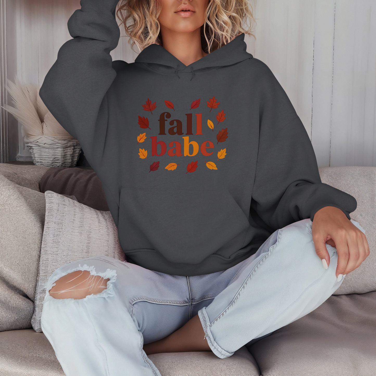 Person wearing a dark gray hoodie with 'fall babe' text and autumn leaves, sitting on a couch.