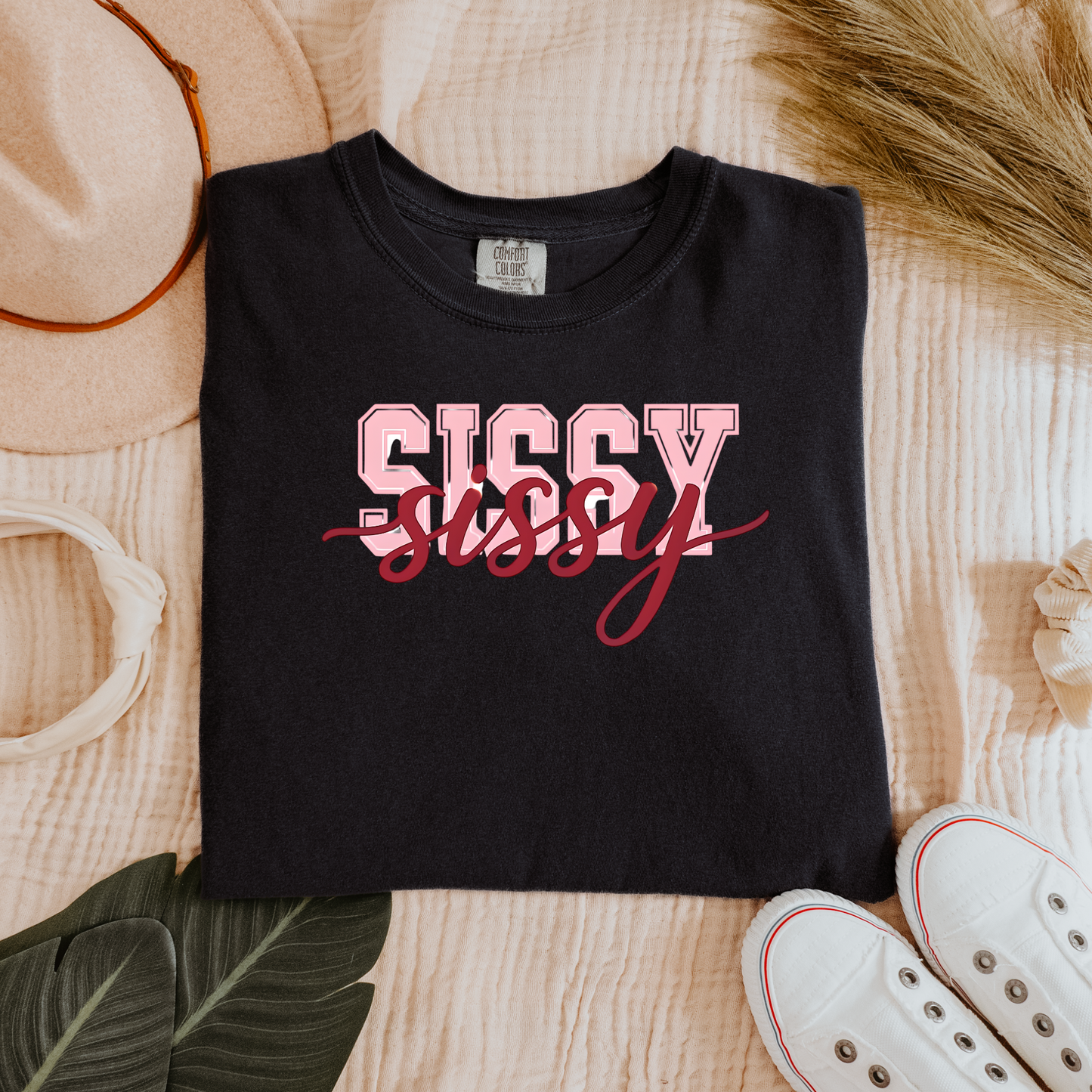 Women wearing t-shirts with the word “SISSY” in pink varsity letters overlaid with dark red cursive script, in colors including charcoal, denim blue, green, black, and ivory, styled in casual everyday scenes