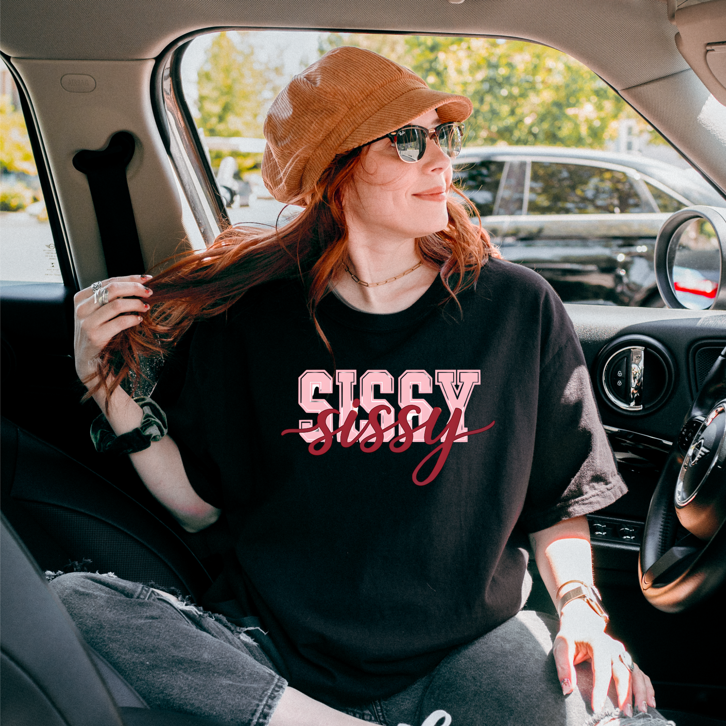 Women wearing t-shirts with the word “SISSY” in pink varsity letters overlaid with dark red cursive script, in colors including charcoal, denim blue, green, black, and ivory, styled in casual everyday scenes