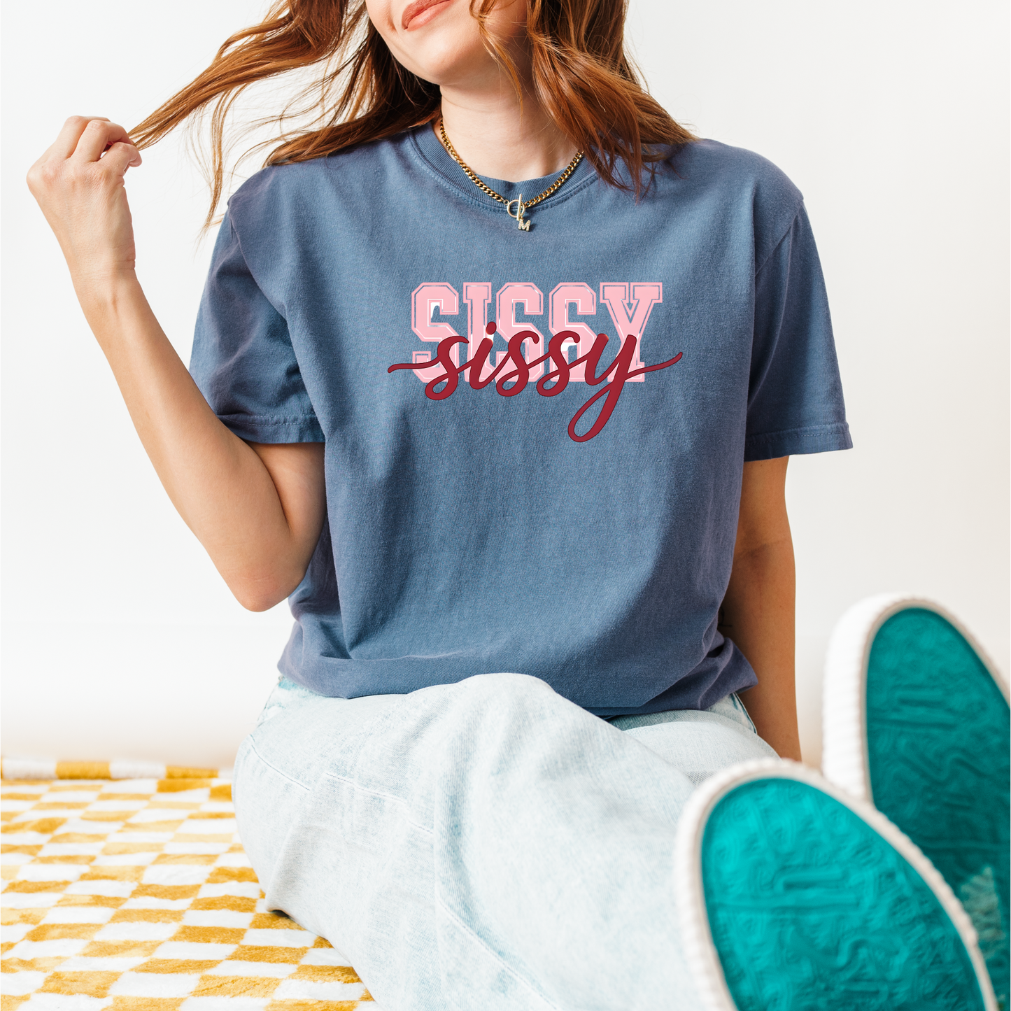 Women wearing t-shirts with the word “SISSY” in pink varsity letters overlaid with dark red cursive script, in colors including charcoal, denim blue, green, black, and ivory, styled in casual everyday scenes