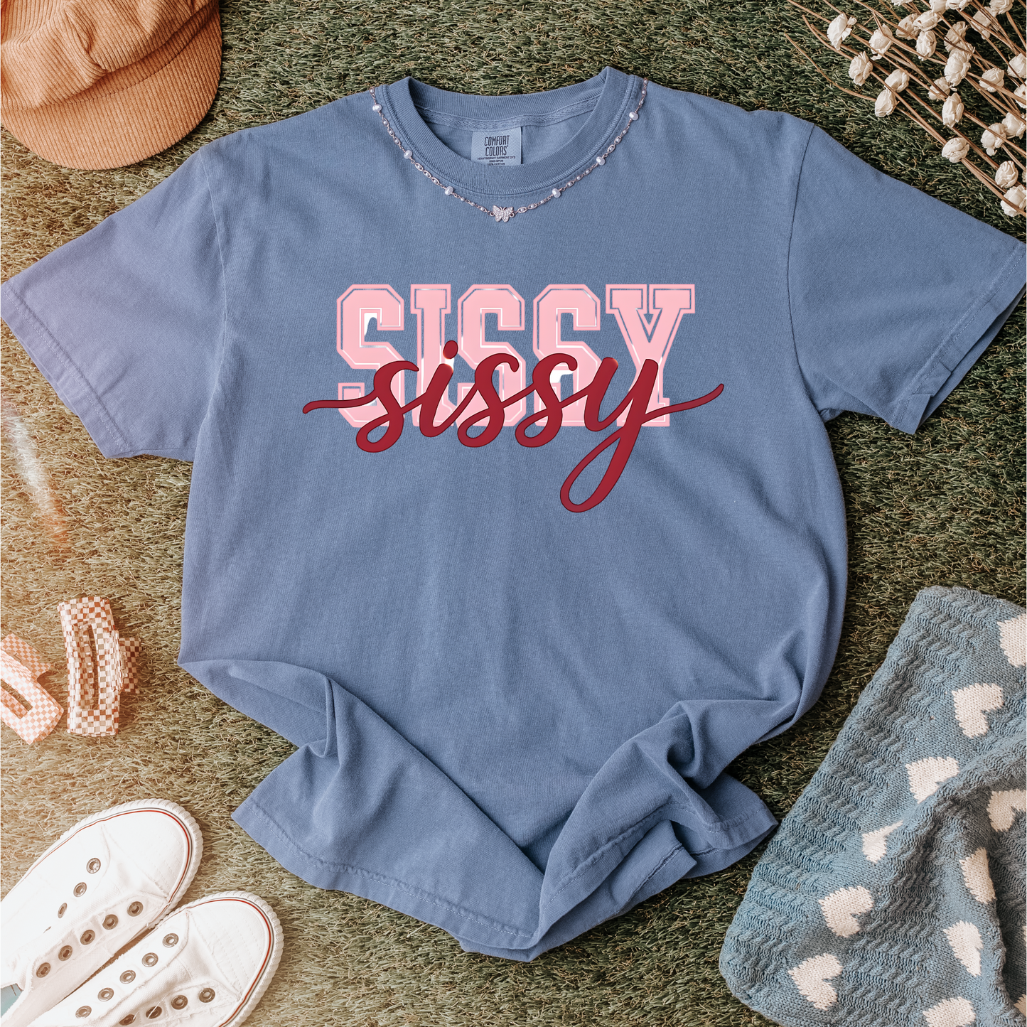 Women wearing t-shirts with the word “SISSY” in pink varsity letters overlaid with dark red cursive script, in colors including charcoal, denim blue, green, black, and ivory, styled in casual everyday scenes