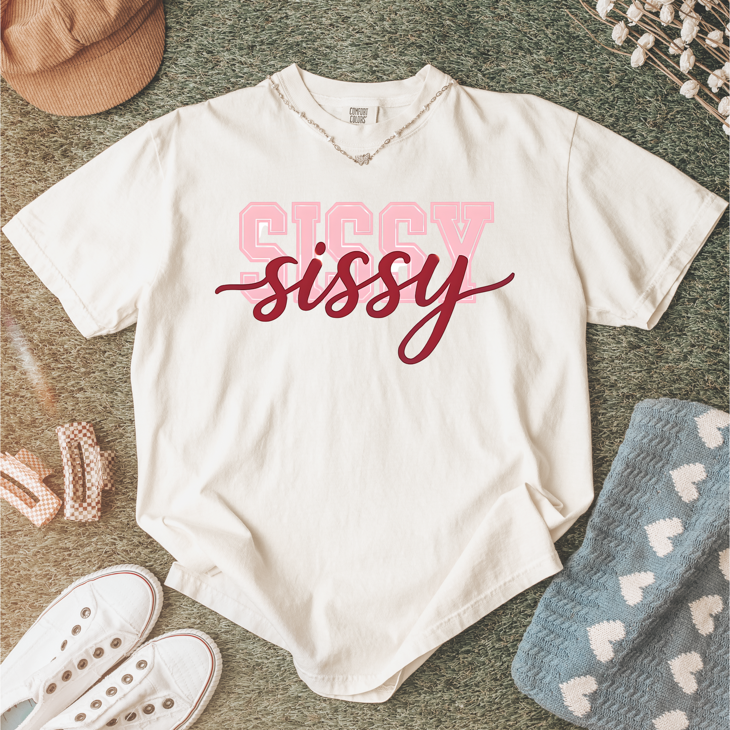 Women wearing t-shirts with the word “SISSY” in pink varsity letters overlaid with dark red cursive script, in colors including charcoal, denim blue, green, black, and ivory, styled in casual everyday scenes