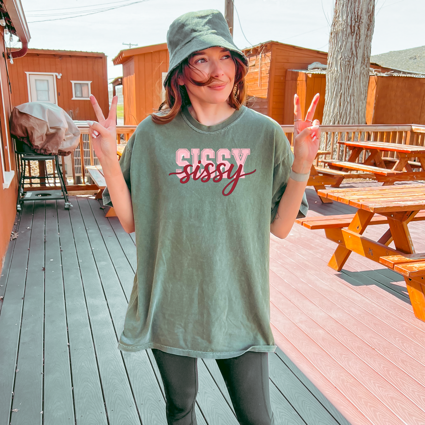 Women wearing t-shirts with the word “SISSY” in pink varsity letters overlaid with dark red cursive script, in colors including charcoal, denim blue, green, black, and ivory, styled in casual everyday scenes