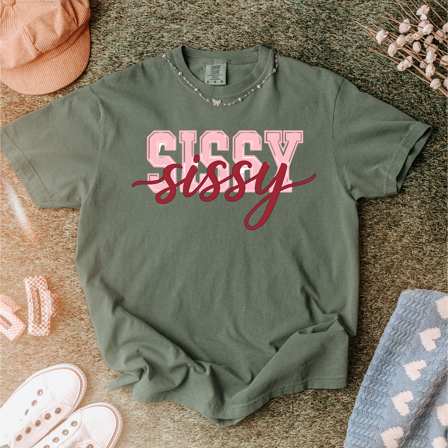 Women wearing t-shirts with the word “SISSY” in pink varsity letters overlaid with dark red cursive script, in colors including charcoal, denim blue, green, black, and ivory, styled in casual everyday scenes