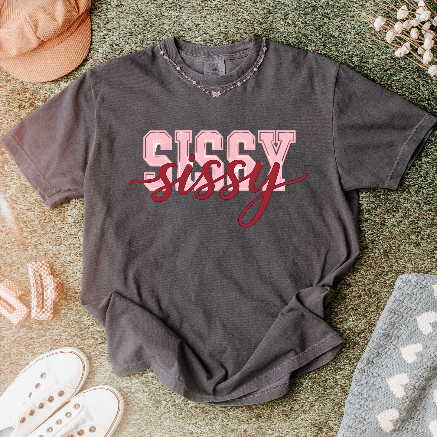 Women wearing t-shirts with the word “SISSY” in pink varsity letters overlaid with dark red cursive script, in colors including charcoal, denim blue, green, black, and ivory, styled in casual everyday scenes