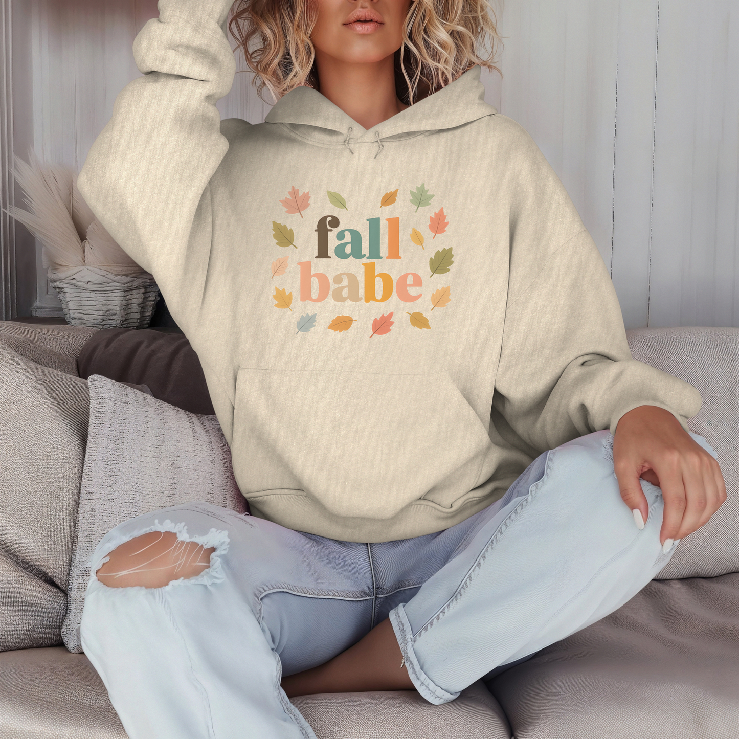 Person wearing a beige hoodie with 'fall babe' text and colorful leaves, sitting on a couch.