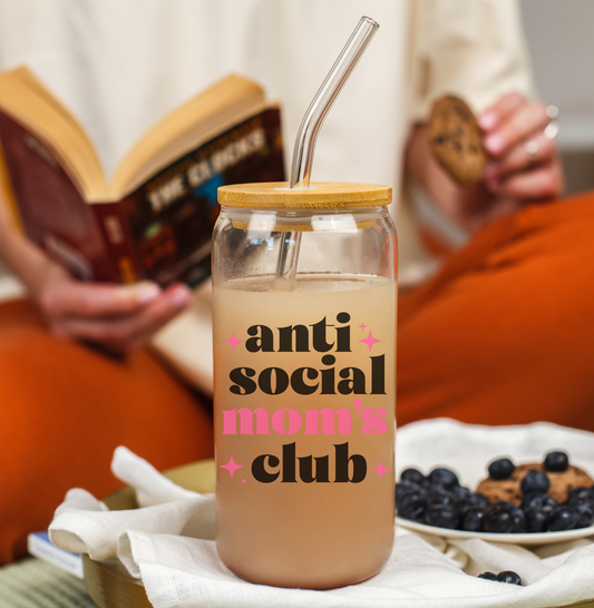 A clear 16 oz glass can tumbler with bamboo lid and glass straw, featuring bold black and pink text reading “Anti-Social Mom’s Club.” The tumbler is styled with beverages and cozy lifestyle scenes.