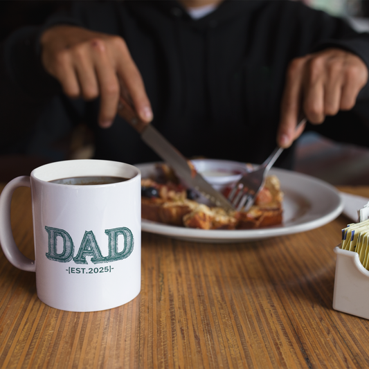 White ceramic coffee mug with bold green vintage-style "DAD" lettering and the text "EST. 2025" below.
