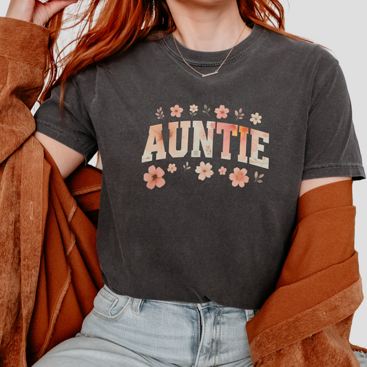 T-shirt featuring the word “AUNTIE” in floral block lettering surrounded by small pastel flowers, shown in rose, lilac, black, slate blue, and charcoal color options