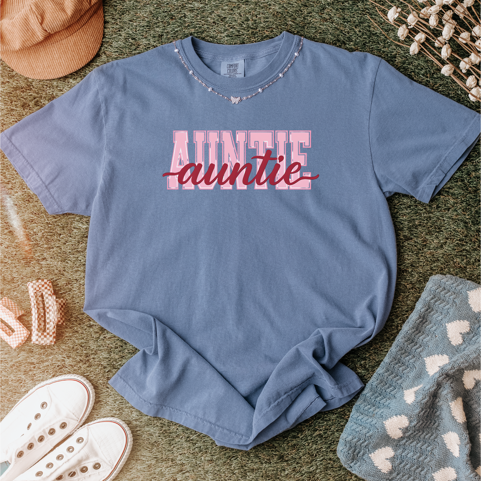 Women wearing graphic t-shirts with the word “AUNTIE” in bold pink varsity letters and a red cursive overlay, available in black, blue, green, and ivory, styled in lifestyle and casual settings