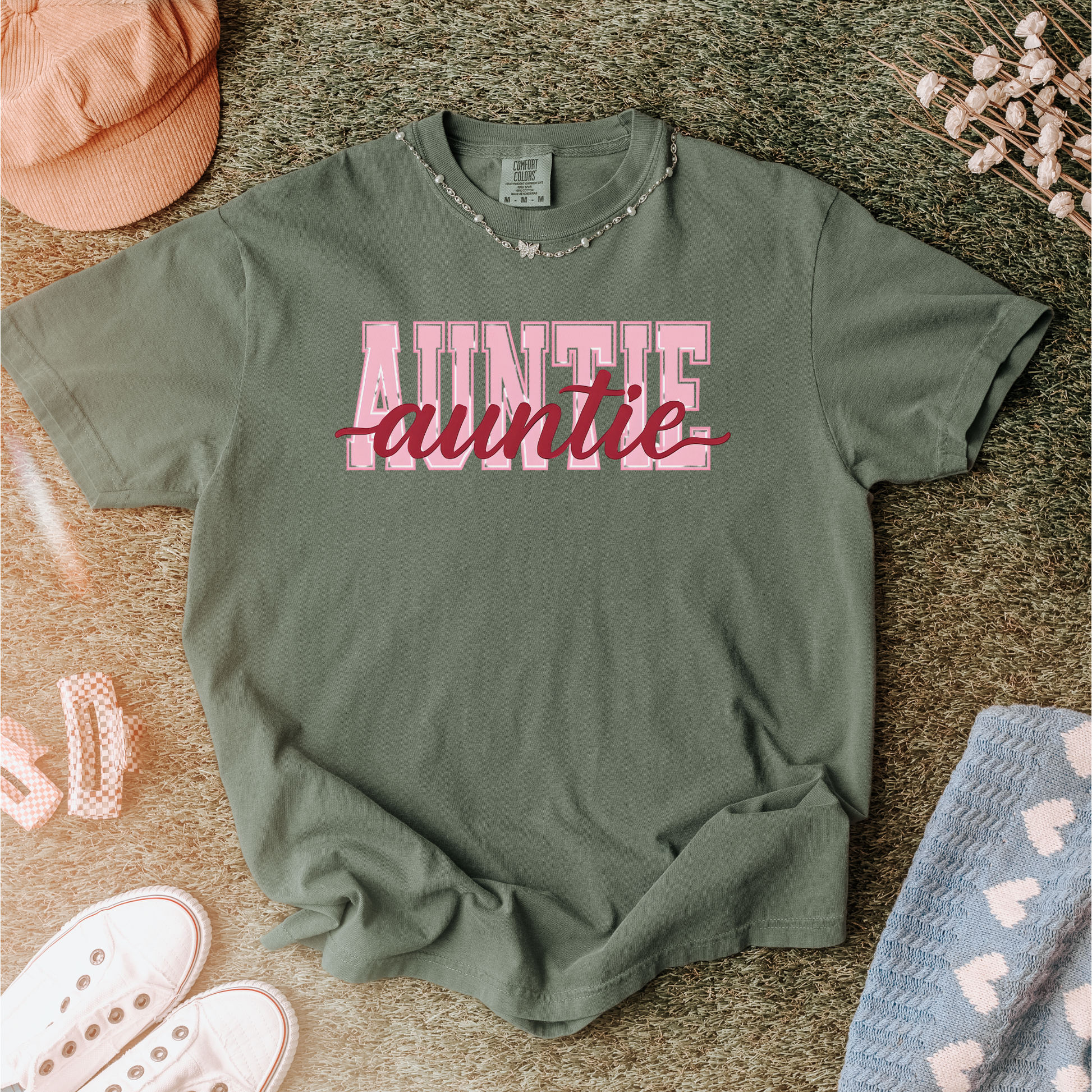 Women wearing graphic t-shirts with the word “AUNTIE” in bold pink varsity letters and a red cursive overlay, available in black, blue, green, and ivory, styled in lifestyle and casual settings
