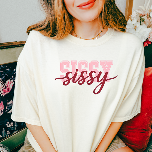 Women wearing t-shirts with the word “SISSY” in pink varsity letters overlaid with dark red cursive script, in colors including charcoal, denim blue, green, black, and ivory, styled in casual everyday scenes